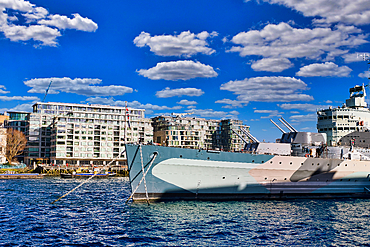 A light-grey warship, moored in a harbor, with modern buildings and a partly cloudy blue sky in the background. Heavy mooring chains are visible in London, UK.