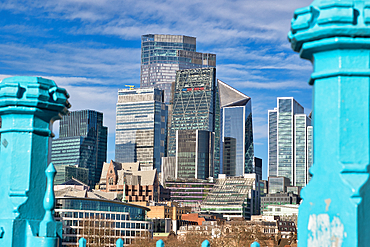 Modern skyscrapers and older buildings in London, viewed through teal bridge railings under a partly cloudy blue sky. The image showcases a mix of architectural styles and contrasts between new and old.