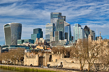 Modern skyscrapers of London's financial district contrast against the historic Tower of London and its surrounding walls under a partly cloudy sky.