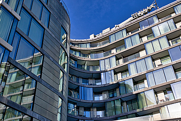 Two modern, multi-story buildings with curved facades and numerous windows and balconies. One building is predominantly gray, the other features glass-fronted balconies. The sky is a clear blue in London, UK.