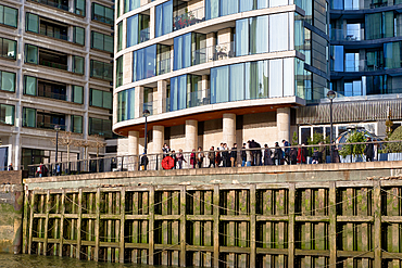 Modern apartment buildings and a group of people stand on a waterfront walkway above a weathered wooden retaining wall. The scene is brightly lit, showcasing architectural details and the people in London, UK.