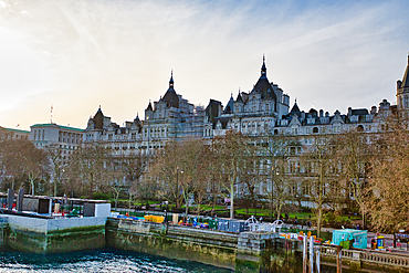 A large, multi-sectioned building with numerous towers and dormers, situated along a waterfront. The building is light-grey stone, partially obscured by bare winter trees. Construction equipment is visible at the water's edge in London, UK.