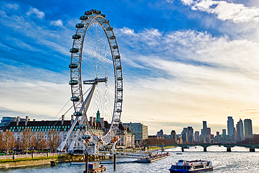 The London Eye Ferris wheel dominates this image, situated on the Thames River. Buildings line the riverbank, and a city skyline is visible in the distance under a partly cloudy sky. Several boats are on the water.