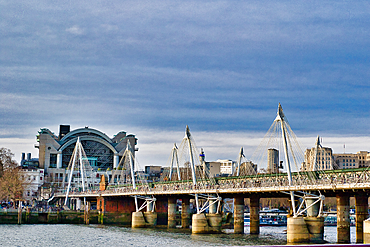 A cable-stayed pedestrian bridge spans a river, with a large modern building in the background under a partly cloudy sky. The scene is dominated by cool tones, with the bridge's white cables and grey structure contrasting against the water and sky in Lon