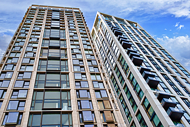 Two modern high-rise residential buildings stand side-by-side against a partly cloudy blue sky. One building features balconies on each floor, while the other has a more recessed window design. Both are light-colored with dark window frames in London,