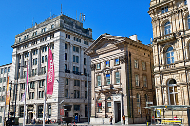 Exterior shot of several buildings, including The National Bank, with people and street furniture, under a bright blue sky in Liverpool, UK.