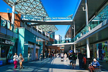 Outdoor shopping center scene featuring shops, people, and elevated walkways under a clear blue sky in Liverpool, UK.
