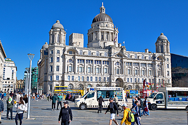 The image shows the Royal Liver Building in Liverpool, England, with people and food trucks in the foreground under a clear blue sky.