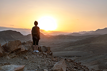 Woman wearing backpack stands alone on the high rock in the dark during sunset and looking at setting sun in the desert mountains of Negev desert in Israel