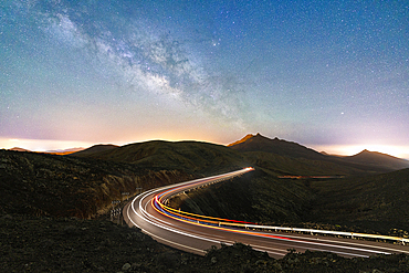 Milky Way glowing over the car trail lights on a winding desert road, Sicasumbre viewpoint, Fuerteventura, Canary Islands, Spain