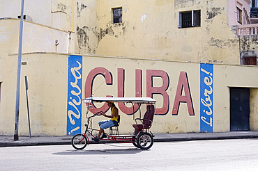 A rickshaw passing a wall sign celebrating Cuban freedom, Havana, Cuba, West Indies, Central America