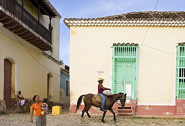 A cowboy riding a horse through the cobbled streets and a woman carrying her baby, Trinidad, Cuba, West Indies, Central America