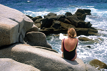 France, Corsica, Ajaccio, Sanguinaires Islands, Woman In Bathing Suit Sitting On Rocks And Reading A Book