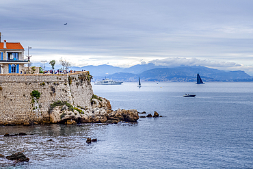 Promenade de Amiral de Grasse, with boats sailing on a calm, overcast day in the Baie des Anges, Antibes, French Riviera. Antibes, French Riviera.