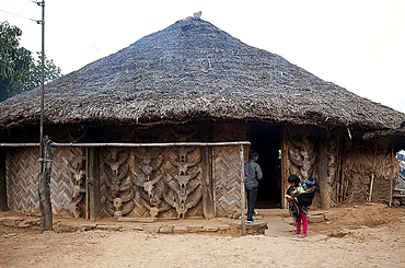 Young girl carrying sibling in cloth papoose outside traditionally constructed palm thatch house, with Mithun skulls, Nagaland, India, Asia