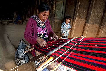 Woman weaving traditional red Naga shawl, young daughter beside her, on the verandah of the family home, Nagaland, India, Asia