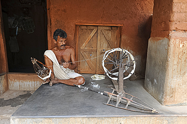 Man on his porch, spinning silk using traditional wooden spinning wheel and stick cages, Nuapatna, Odisha, India, Asia