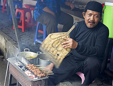 Muslim man in the market cooking chicken satay, fanning the charcoal with a palm leaf fan, Solo, Java, Indonesia, Southeast Asia, Asia