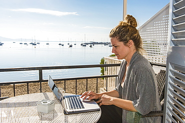 Woman working on her laptop on a balcony overlooking the ocean, Port de Pollenca, Mallorca, Balearic Islands, Spain, Mediterranean, Europe 