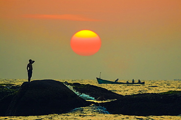 Man on intertidal rocks and fishing boat at sunset on beautiful unspoilt Kizhunna Beach, south of Kannur, Kannur, Kerala, India, Asia