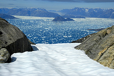 Wide open view over a snow field through rocks to icebergs in Sermilik Fjord Eastgreenland