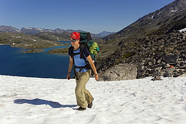 Young woman hiking, backpacking, hiker with backpack, snow field, descending towards summit of historic Chilkoot Trail, Chilkoot Pass, Crater Lake behind, alpine tundra, Yukon Territory, British Columbia, B. C., Canada