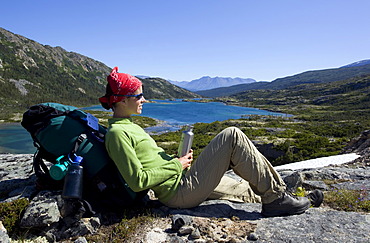 Young woman resting, hiker with backpack, water bottle, panorama, deep lake behind, historic Chilkoot Trail, Chilkoot Pass, alpine tundra, Yukon Territory, British Columbia, B. C., Canada