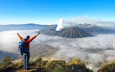 Young man with raised arms in front of volcanic landscape, view in Tengger Caldera, smoking volcano Gunung Bromo, in front Mt. Batok, in the back Mt. Kursi, Mt. Gunung Semeru, National Park Bromo-Tengger-Semeru, Java, Indonesia, Asia