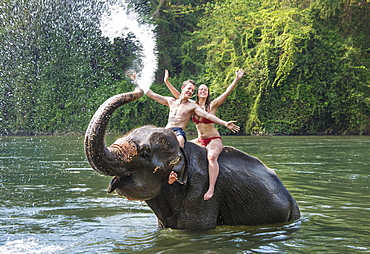 Two teenagers sitting on an Asian Elephant (Elephas maximus), being sprayed with water from the trunk, Kanchanaburi, Thailand, Asia