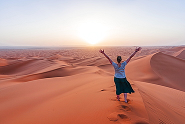 Female hiker is stretching arms in the air, red sand dune in the desert, sunset, Erg Chebbi, Merzouga, Sahara, Morocco, Africa