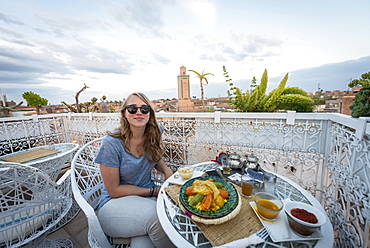 Young woman on a roof terrace in the restaurant, Moroccan food, tagine, view of the old town, mosque with minaret, evening mood, Marrakech, Morocco, Africa