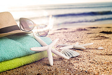 Summer accessories as colorful towels, straw hat, sun glasses and shells on the sandy beach