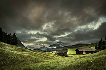 Small huts on mountain meadow with dramatic cloud sky and Wetterstein mountains in the background, Mittenwald, Bavaria, Germany, Europe