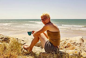 Woman, sitting on rock by the sea and drinking a cup of coffee, camping, Portbail, Normandy, France, Europe