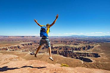 Young man jumping with outstretched arms, view from Grand Viewpoint, rugged gorges of the Green River, canyon landscape, erosion landscape, rock formations, Monument Basin, White Rim, Island in the Sky, Canyonlands National Park, Utah, USA, North America