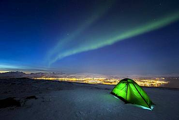 Green illuminated tent in the snow and aurora borealis above Fjellheisen, Tromsoe, Norway, Europe