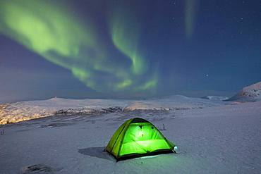 Green illuminated tent in the snow and aurora borealis above Fjellheisen, Tromsoe, Norway, Europe