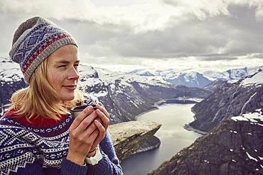 Young woman in Norwegian sweater drinks tea, enjoys view over Trolltunga, Norway, Europe