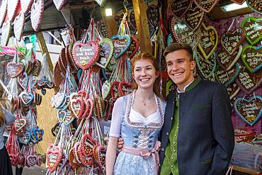 Young couple in traditional traditional traditional traditional traditional traditional traditional traditional traditional traditional costume in front of gingerbread hearts, gingerbread stand, Wiesn, Wiesn, Oktoberfest, Munich, Upper Bavaria, Bavaria, Germany, Europe