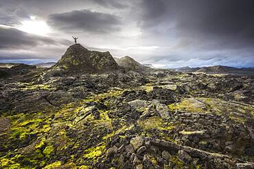 Man on a hill in a black lava field with green moss stretching hands into the dramatic sky, Leihrnjukur in the Krafla, Skutustaoir, Norourland eystra, Iceland, Europe