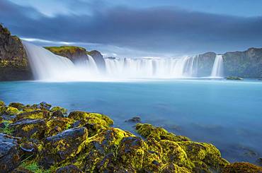 Long time exposure of a turquoise waterfall in volcanic landscape with dramatic clouds and green moss on rocks, Godafoss, Pingeyjarsveit, Norourland eystra, Iceland, Europe