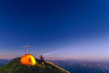 Cross of the Kreuzspitze by a starry sky with tent and mountaineer at the summit, in the background the Lechtal Alps, Elmen, Lechtal Alps, Ausserfern, Tyrol, Austria, Europe