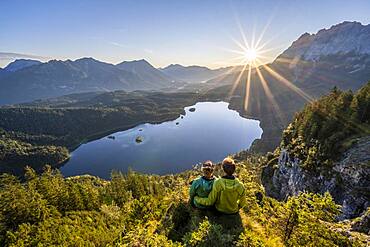 Two (hikers) looking at Eibsee lake at sunrise, sun shining over Bavarian alpine foothills, right Zugspitze, Wetterstein mountains near Grainau, Upper Bavaria, Bavaria, Germany, Europe