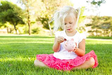 Cute little girl having fun with her piggy bank outside on the grass