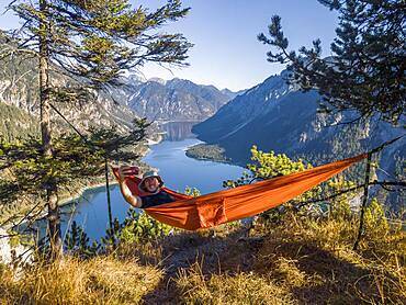 Man taking pictures of himself with mobile phone, hiker sitting in hammock with view of mountains with lake, Plansee, Ammergau Alps, district Reutte, Tyrol, Austria, Europe