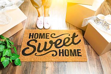 Woman in pink shoes and sweats standing near home sweet home welcome mat, boxes and plant