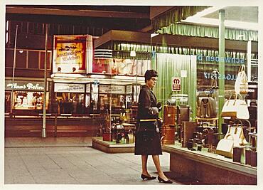 Stuttgart at night in 1963: A young woman with a coat and handbag looks at the display of handbags and suitcases in a leather goods shop, in the back a cinema with the announcement of the film Ein fast anstaendiges Maedchen starring Liselotte Pulver, Baden-Wuerttemberg, Germany, Europe