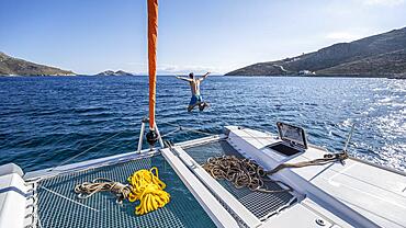 Young man jumps into the water, ropes on a sailing catamaran, sailing trip, Tilos, Dodecanese, Greece, Europe