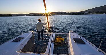 Young man standing by the foresail, sunset with sun star, Sailing a sailing catamaran, Dodecanese, Greece, Europe