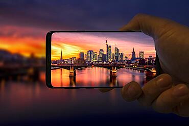 Mobile phone takes photo, A hand holds a mobile phone in the picture, on it to see, The old bridge over the Main with skyline, high-rise buildings in the banking district in the evening, sunset, Frankfurt am Main, Hesse, Germany, Europe